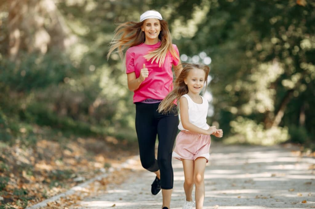 Smiling, a woman in pink runs with a young girl on a leaf-strewn park path